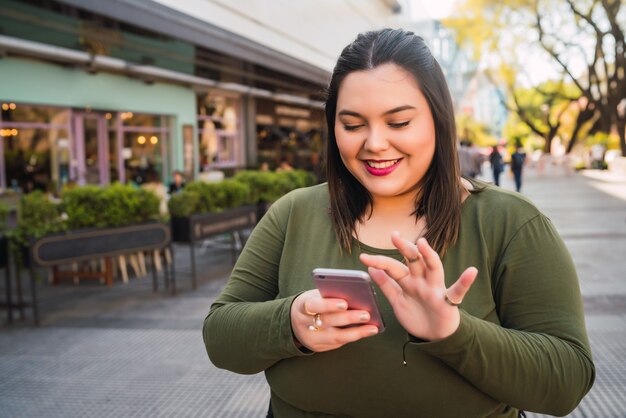 Retrato de mujer joven de talla grande escribiendo un mensaje de texto en su teléfono móvil al aire libre en la calle. Concepto de tecnología.