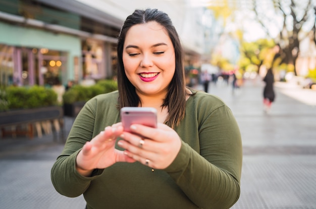 Retrato de mujer joven de talla grande escribiendo un mensaje de texto en su teléfono móvil al aire libre en la calle. Concepto de tecnología.