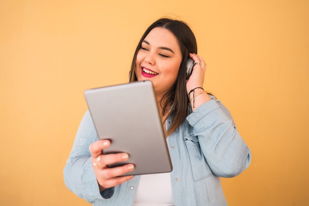 Retrato de mujer joven de talla grande escuchando música con auriculares y tableta digital al aire libre sobre fondo amarillo.