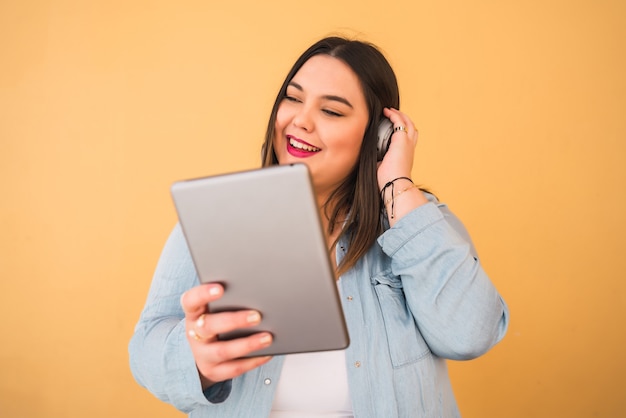 Retrato de mujer joven de talla grande escuchando música con auriculares y tableta digital al aire libre sobre fondo amarillo.