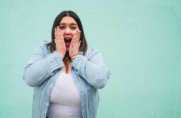 Retrato de mujer joven de tamaño pluse con una expresión de asombro mientras está de pie contra la pared azul claro al aire libre.