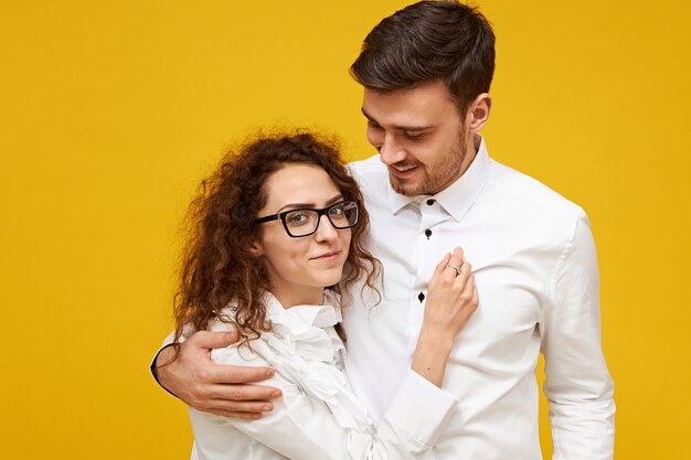 Retrato de mujer joven tímida con cabello oscuro y rizado con sonrisa tímida abrazando a su alto novio con rastrojo. Chico guapo va a besar a su encantadora novia, estar enamorado