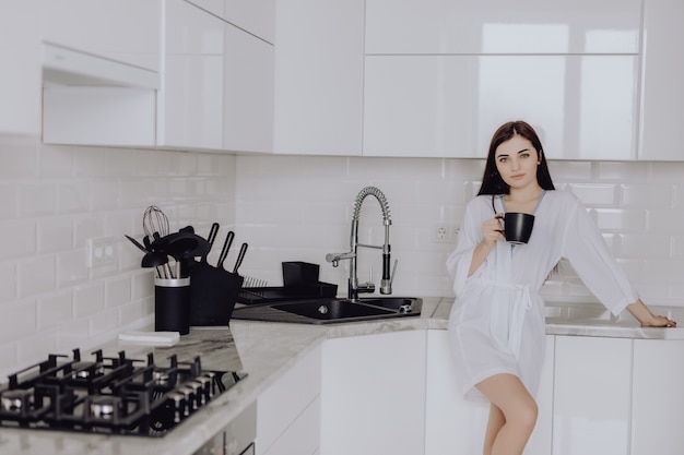 El retrato de la mujer joven en el traje blanco después del baño sostiene una taza con café o té contra la pared de la cocina. ciérrese encima de la sonrisa dentuda de la cara.