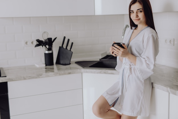 El retrato de la mujer joven en el traje blanco después del baño sostiene una taza con café o té contra la pared de la cocina.