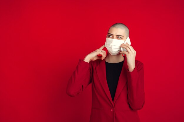 Retrato de mujer joven en traje rojo y mascarilla blanca hablando por teléfono aislado en estudio rojo