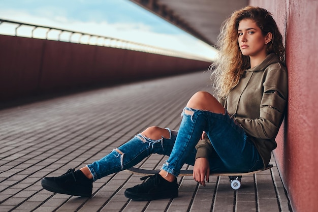 Retrato de una mujer joven vestida con una sudadera con capucha y jeans rotos apoyados en una pared mientras se sienta en una patineta en la acera del puente.