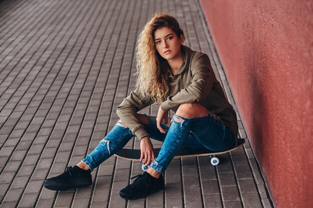 Retrato de una mujer joven vestida con una sudadera con capucha y jeans rotos sentada en una patineta en un puente peatonal.