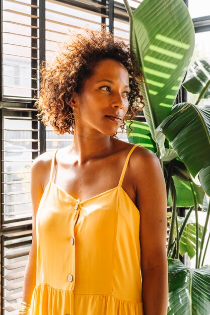 Retrato de mujer joven en vestido amarillo de pie cerca de la ventana