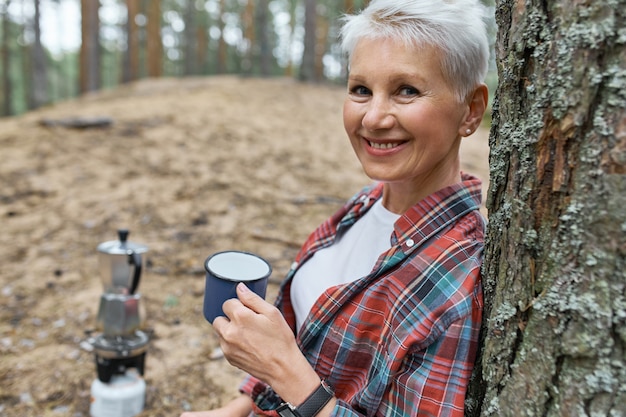 Foto gratuita retrato de mujer jubilada enérgica recostada sobre pino sosteniendo una taza, bebiendo té ella del agua que ella hirvió en la tetera en el quemador de camping