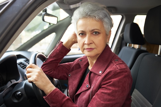 Retrato de mujer jubilada seria con corte de pelo corto sentado dentro del coche, pasando el examen de conducir, sintiéndose nervioso.
