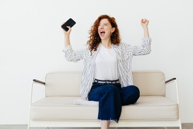 Retrato de mujer l sosteniendo y usando su teléfono celular sacudiendo el puño gritando sí celebrando la victoria o la victoria jugando juegos en línea leyendo buenas noticias sentada en el sofá