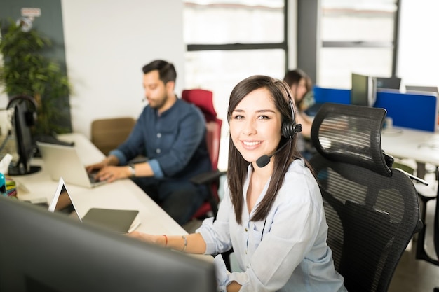 Retrato de una mujer latina bastante joven que lleva un auricular sentado en su escritorio de oficina con compañeros de trabajo en segundo plano.