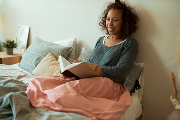 Retrato de mujer latina joven feliz con cabello castaño rizado relajándose en casa, sentada en la cama con libro abierto, disfrutando de la lectura