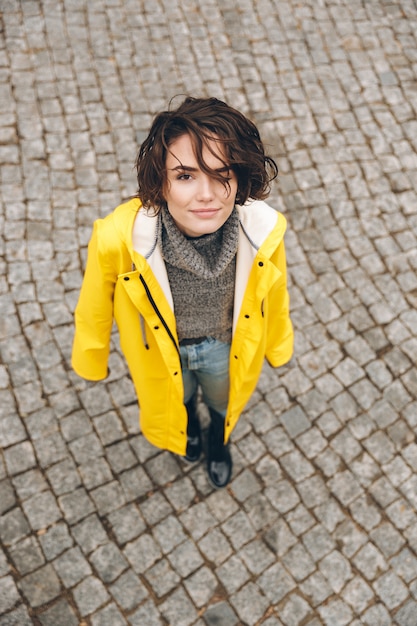 Retrato de mujer linda en elegante abrigo amarillo mirando hacia la cámara mientras está de pie sobre adoquines durante su caminata