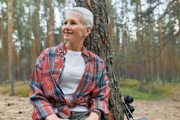 Retrato de mujer madura feliz con har rubio corto sentado bajo un árbol en camisa a cuadros mirando a su alrededor, admirando el hermoso bosque de pinos, habiendo relajado la expresión facial, sonriendo. Senderismo y naturaleza