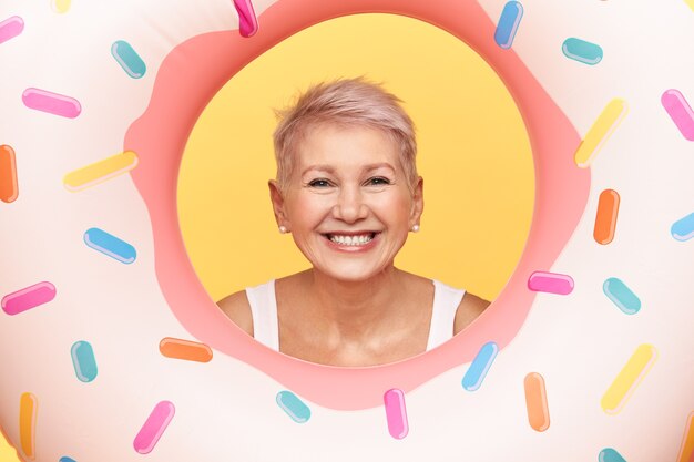 Retrato de mujer madura positiva feliz con peinado corto asomando la cabeza en el agujero de rosquilla rosa inflable, divirtiéndose en la playa, nadando, sonriendo ampliamente.