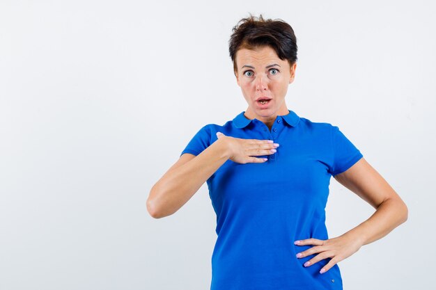 Retrato de mujer madura sosteniendo la mano en el pecho en forma de cuestionamiento en camiseta azul y mirando perplejo vista frontal