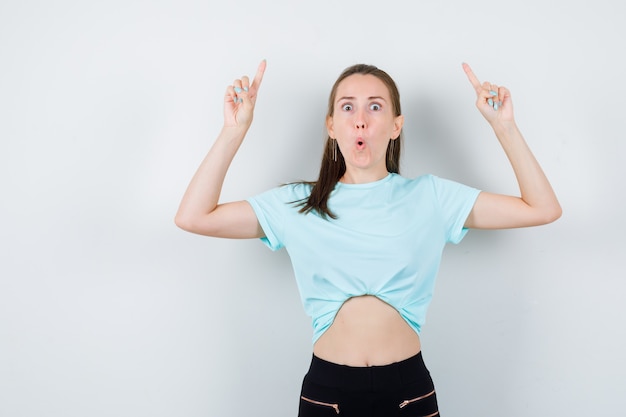 Retrato de mujer maravillosa apuntando hacia arriba en blusa, pantalones y mirando sorprendido vista frontal