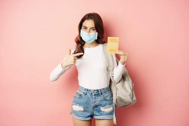 Retrato de mujer con mascarilla médica que viaja con certificación de vacunación internacional covid, sosteniendo mochila, de pie sobre fondo rosa.