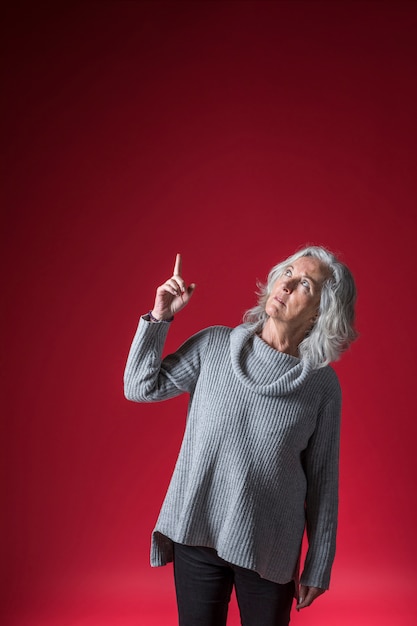 Foto gratuita retrato de una mujer mayor apuntando el dedo hacia arriba contra un fondo rojo brillante