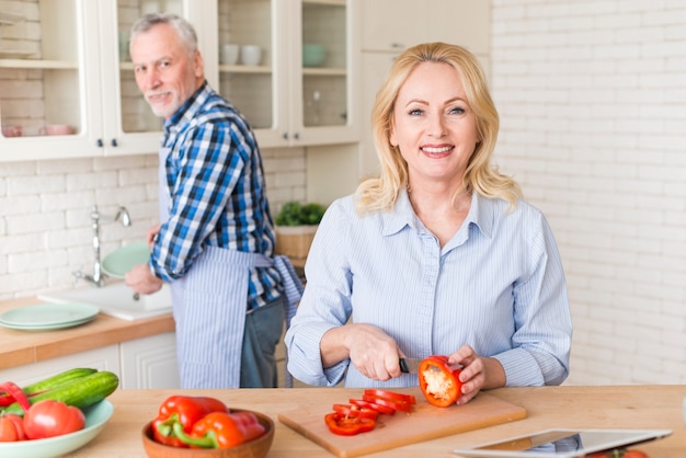 Retrato de una mujer mayor cortando el pimiento con un cuchillo y su esposo lavando platos en el fregadero de la cocina