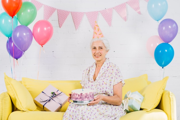 Retrato de una mujer mayor feliz que se sienta en el sofá con la torta de cumpleaños