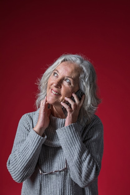 Retrato de una mujer mayor que habla en el teléfono móvil que mira para arriba contra fondo rojo
