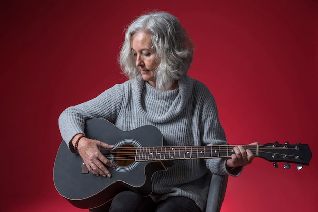 Retrato de una mujer mayor que se sienta en la silla que toca la guitarra contra fondo rojo