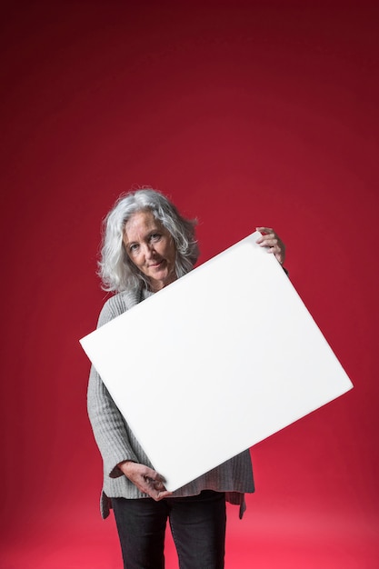 Retrato de una mujer mayor que sostiene el cartel blanco a disposición contra el fondo rojo