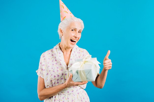 Retrato de una mujer mayor con el regalo de cumpleaños que gesticula los pulgares para arriba en el contexto azul