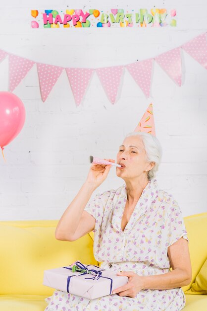 Retrato de una mujer mayor con el regalo de cumpleaños que se sienta en el sofá que sopla el cuerno del partido