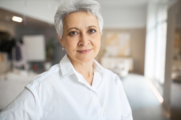 Retrato de mujer de mediana edad segura seria con pelo corto gris, ojos verdes, arrugas y sonrisa encantadora posando en interiores con los brazos cruzados