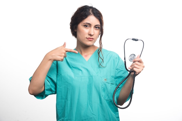 Retrato de mujer médico apuntando con el dedo en el estetoscopio sobre fondo blanco.