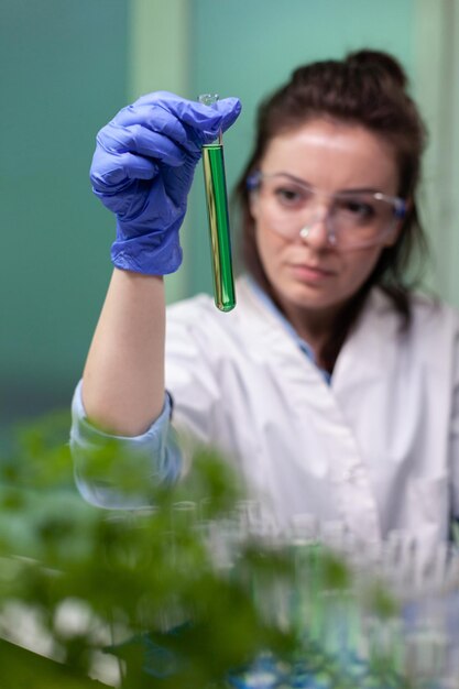 Retrato de mujer médico bioquímico sosteniendo un tubo de ensayo médico con líquido verde analizando la sustancia genética durante el experimento biológico. Médico científico que trabaja en el laboratorio farmacéutico
