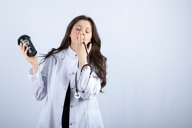 Retrato de mujer médico con estetoscopio y taza de café durmiendo en la pared blanca.