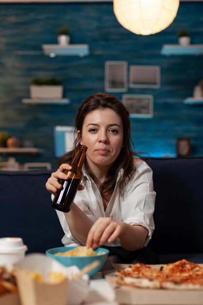 Retrato de mujer mirando a cámara sosteniendo una botella de cerveza y comiendo papas fritas del tazón en la mesa con pizza grande y cena para llevar. Trabajador de oficina en el sofá relajante después del menú para llevar.