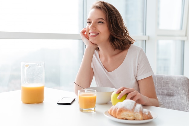 Retrato de mujer morena alegre y soñadora sosteniendo su cabeza mientras desayunando en la mesa de la cocina