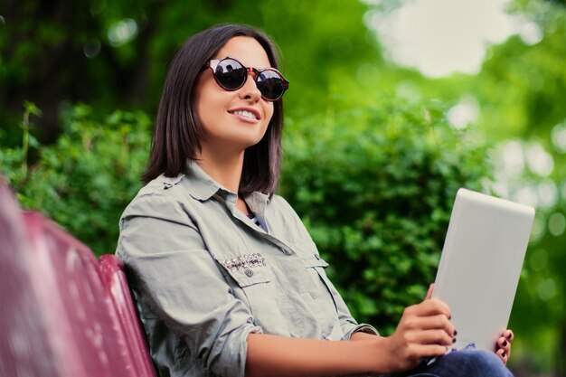 Retrato de mujer morena atractiva se sienta en un banco y tiene tablet PC en un parque de verano.