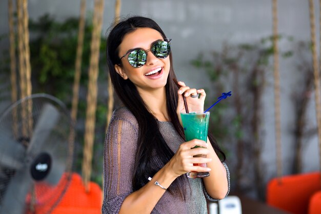 Retrato de mujer morena bonita feliz bebiendo cóctel frío sabroso, traje elegante y gafas de sol con espejo de cerca, disfruta de su fin de semana, tiempo de fiesta.