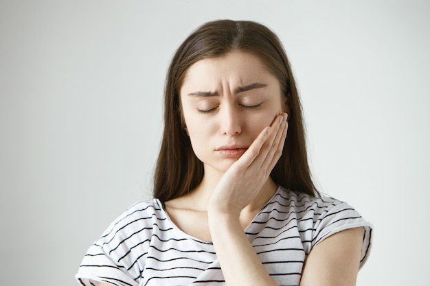 Foto gratuita retrato de mujer morena caucásica joven deprimida infeliz cerrando los ojos con frustración y presionando la mano contra la mejilla, tratando de calmar el dolor mientras sufre de dolor de muelas, con caries dental