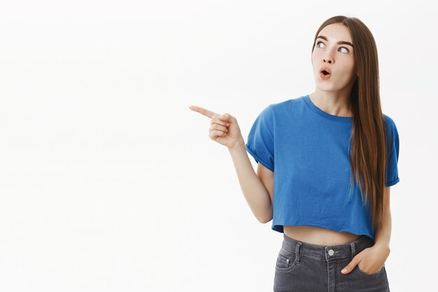 Retrato de mujer morena encantadora impresionada y asombrada con cabello largo y saludable en camiseta azul doblando los labios con asombro apuntando y mirando sorprendido a la izquierda