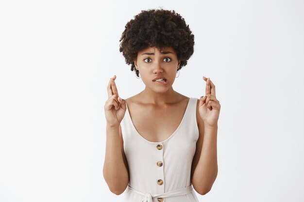 Retrato de mujer morena encantadora preocupada intensa con peinado afro, mordiendo el labio nerviosamente, frunciendo el ceño y mirando ansiosamente mientras cruza los dedos con esperanza