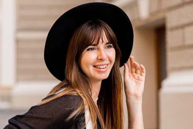 Retrato de mujer morena con estilo en traje casual y sombrero negro de cerca. Disfrutando de las vacaciones en la ciudad europea