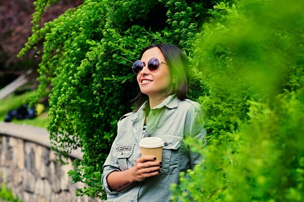 Retrato de mujer morena con gafas de sol bebe café mientras viaja en un parque de verano.