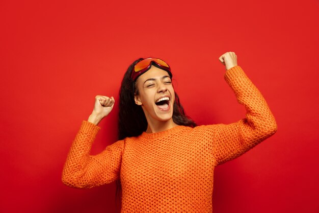 Retrato de mujer morena joven afroamericana en pasamontañas sobre fondo rojo de estudio. Concepto de emociones humanas, expresión facial, ventas, publicidad, deportes de invierno y vacaciones. Celebre como ganador.