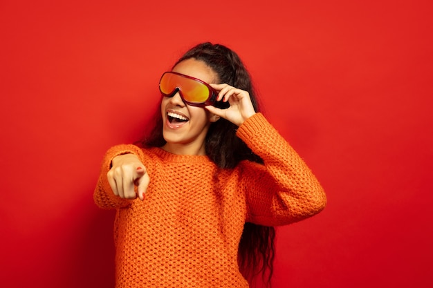 Retrato de mujer morena joven afroamericana en pasamontañas sobre fondo rojo de estudio. Concepto de emociones humanas, expresión facial, ventas, publicidad, deportes de invierno y vacaciones. Sonriendo, señalando.