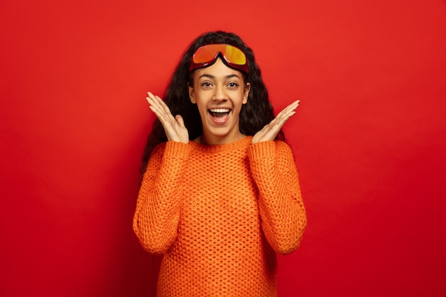 Retrato de mujer morena joven afroamericana en pasamontañas sobre fondo rojo de estudio. Concepto de emociones humanas, expresión facial, ventas, publicidad, deportes de invierno y vacaciones. Sorprendido, asombrado.