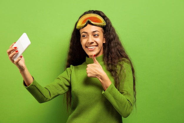 Retrato de mujer morena joven afroamericana en pasamontañas sobre fondo verde de estudio. Concepto de emociones humanas, expresión facial, ventas, publicidad, deportes de invierno y vacaciones. Haciendo selfie o vlog.