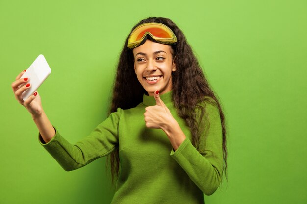 Retrato de mujer morena joven afroamericana en pasamontañas sobre fondo verde de estudio. Concepto de emociones humanas, expresión facial, ventas, publicidad, deportes de invierno y vacaciones. Haciendo selfie o vlog.