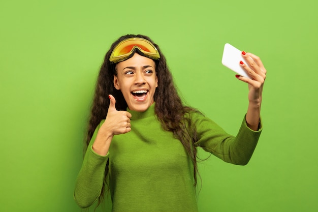 Retrato de mujer morena joven afroamericana en pasamontañas sobre fondo verde de estudio. Concepto de emociones humanas, expresión facial, ventas, publicidad, deportes de invierno y vacaciones. Haciendo selfie o vlog.